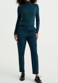 Pull à col roulé bleu sarcelle et pantalon ajusté assorti, avec une fermeture par bouton, une texture lisse et une coupe slim. Des chaussures noires à lacets complètent le look.