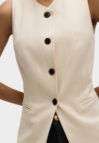 Creme vest med v-udskæring, med fem sorte knapper, sidelommer og et slankt design. Glat tekstur og skræddersyet pasform.