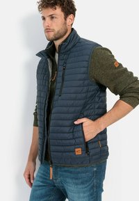 Marineblauer gesteppter Weste mit hohem Kragen, mit Reißverschlusstaschen und subtilen orangefarbenen Akzenten, über einem grünen strukturierten Pullover getragen.