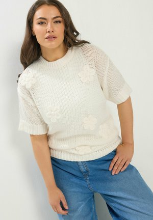 Frau trägt ein weißes Strickpullover mit kurzen Ärmeln und floralen Mustern, kombiniert mit locker sitzenden blauen Jeans, posiert vor einer weißen Wand.