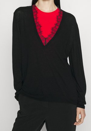 Personne portant un pull noir en V superposé à un haut rouge à col volanté, et un pantalon noir sur un fond uni.