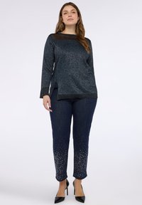 Maglione a maglia metallizzata blu scuro con pannello in rete sul petto, abbinato a jeans in denim scuri con dettagli in metallo argentato sull'orlo.