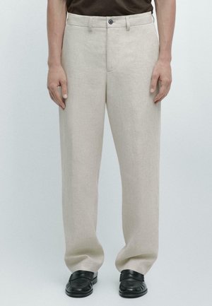 Mann trägt beige gerade geschnittene Hose mit Knopf- und Reißverschluss, kombiniert mit schwarzen Slippern, gezeigt von der Taille bis zu den Füßen vor einfachem Hintergrund.