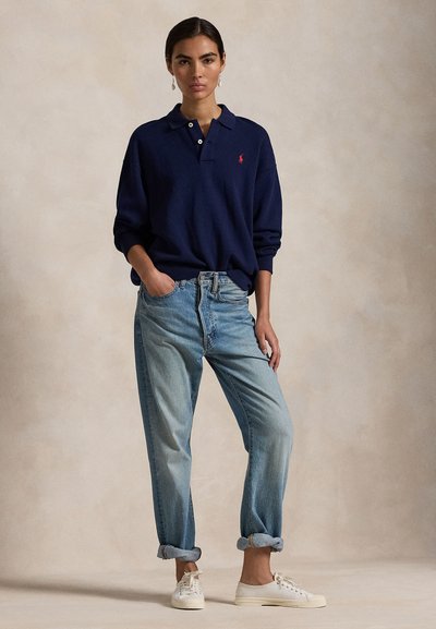 Naine seisab ühe käega taskus, kannab tumesinist pika varrukaga polot, helesiniseid ülesrullitud teksapükse, valgeid tossusid ja ripatavaid kõrvarõngaid.