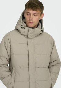 Beige Pufferjacke mit hohem Kragen, gepolsterten Bereichen und einer verstellbaren Kapuze. Verfügt über Fronttaschen und eine glatte Textur.