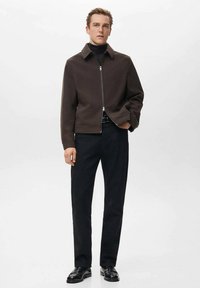 Brązowa kurtka na zamek z kołnierzem, zestawiona z czarnymi spodniami i butami, na modelu na jednolitym białym tle.