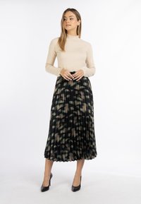 Sort plisseret nederdel med et abstrakt grønt og brunt mønster, parret med en beige langærmet top. Sorte højhælede sko fuldender looket.