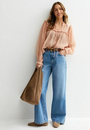 Vrouw die een perzikkleurige blouse en wijde blauwe jeans draagt, een grote bruine draagtas vasthoudt, glimlacht en tegen een witte muur staat.