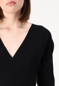 Top preto de malha canelada com decote em V, mangas longas e design envolvente. Textura suave com linhas horizontais subtis.