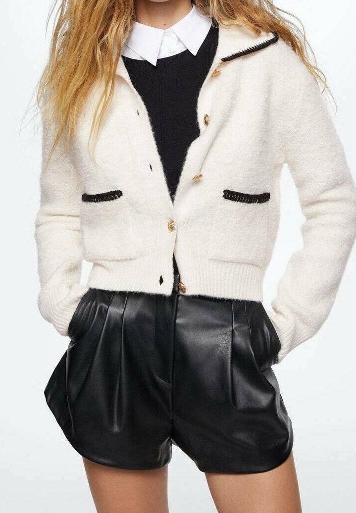 Krémově zbarvený pletený kardigan s černými akcenty, s žebrovaným lemem, zlatými knoflíky a dvěma předními kapsami, zkombinovaný s černými koženými kraťasy.