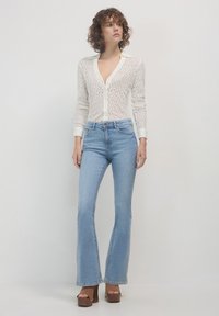 Cardigan bianco in pizzo a maniche lunghe con scollatura a V e chiusura con bottoni, abbinato a jeans a campana azzurri chiari e décolleté con piattaforma marroni.