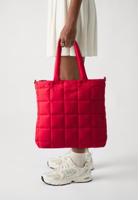 Sac fourre-tout matelassé rouge avec texture rembourrée, deux poignées et fermeture éclair. Porté avec des baskets blanches et une robe de couleur claire.