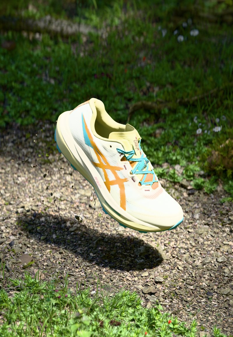 Ljust gult sportskor med orange design, blå snören och texturerad sula, svävande över en grusytan med grönt gräs.
