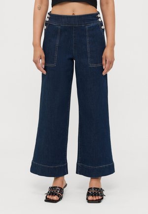 Jean bleu foncé à jambes larges avec des détails de boutons à la taille, porté avec des sandales noires ouvertes cloutées sur une personne debout.