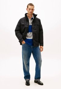 Svart isolerad jacka med hög krage, två framfickor, bärs över en blå, grå och vit randig skjorta och jeans. Svarta skor.