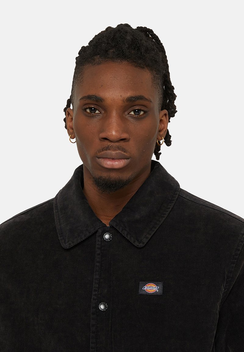 Veste en velours côtelé noir avec un col évasé, fermeture à boutons et un petit patch logo sur la poitrine. Comprend un tissu texturé et en relief.