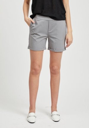 Mujer con pantalones cortos grises con dobladillo enrollado, top negro sin mangas y zapatos blancos tipo slip-on con tachuelas plateadas, de pie frente a un fondo liso.