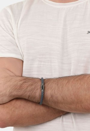 Bracciale in pelle intrecciata grigia con chiusura metallica, indossato su un avambraccio. Lo sfondo presenta una camicia chiara e sottile.