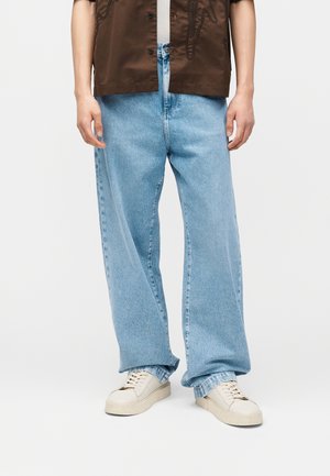 Person iført lyseblå vidde jeans, off-white sneakers, en hvid undertrøje og en brun kortærmet skjorte, stående mod en ensfarvet baggrund.