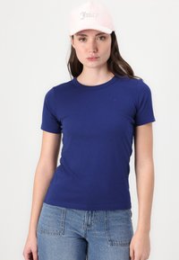 Temno modra oprijeta majica s kratkimi rokavi in okroglo izrezom, v kombinaciji s svetlo modrimi jeans hlačami. Roza kapa z napisom "Juicy" iz kamnov.