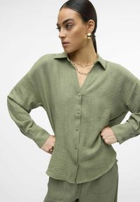 Camicia verde oliva, con bottoni, realizzata in un tessuto testurizzato. Presenta un colletto a V, maniche lunghe e una vestibilità comoda. Include un colletto a punta e polsini con bottoni.