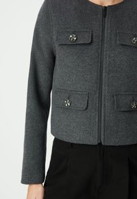 Szary płaszcz z mieszanki wełny z zamkiem błyskawicznym, dwoma przednimi kieszeniami z dekoracyjnymi guzikami i długimi rękawami. Tekstura jest miękka i lekko usztywniona.