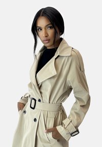 Beige trenchcoat med glansig finish, stora slag, dubbelknäppta knappar och ett svart spännband. Har sidofickor och manschettremmar.