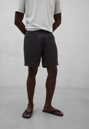 Mann trägt weißes T-Shirt, dunkle knielange Shorts mit geknöpften Gesäßtaschen und schwarze Flip-Flops, steht auf grauem Boden.