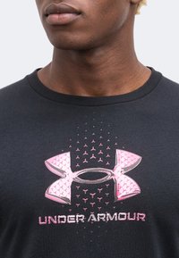 Νεαρός άνδρας με μαύρο πουκάμισο Under Armour με μεγάλο ροζ λογότυπο και όνομα μάρκας στο κέντρο του στήθους.