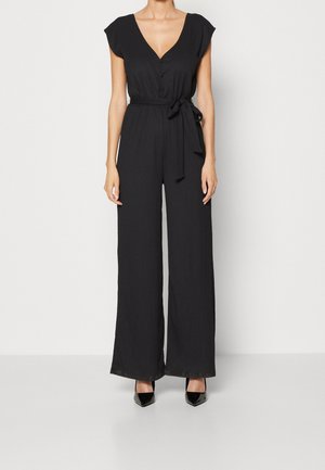 Kvinde iført sort jumpsuit med korte ærmer, V-hals, knapper, bælte bundet i taljen, bukser med vide ben og sorte spidse hæle.