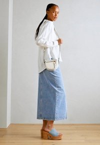 Camisa ligera blanca de manga larga, falda de mezclilla azul de cintura alta y zuecos azules de plataforma con tacones de corcho. Bolso cruzado de color claro.