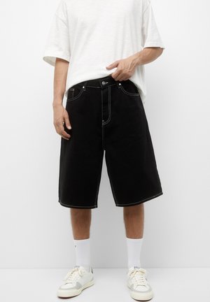 Schwarze Denim-Shorts mit weißen Nähten, knielang und in lockerer Passform. Mit Vordertaschen und einem Knopfverschluss am Bund.