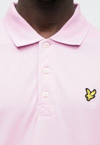 Rožinė polo marškinėliai su klasikinio stiliaus apykakle, dviem sagomis ir kairėje krūtinės pusėje esančiu juodo ir geltono siuvinėto paukščio logotipu. Audinys turi tekstūrą.