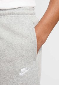Pantalon de jogging gris Nike avec taille élastique, poches latérales et logo blanc brodé sur la cuisse gauche. Texture de tissu douce.