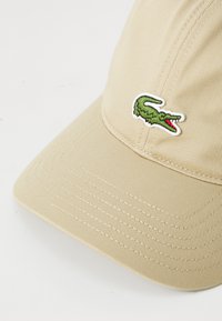 Beige keps i texturerat tyg, med en grön broderad krokodil-logotyp framtill. Böjd skärm och synliga sömdetaljer.