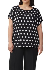 Vrouw in een zwart t-shirt met korte mouwen met onregelmatige witte stippen, gecombineerd met zwarte wijde broek.