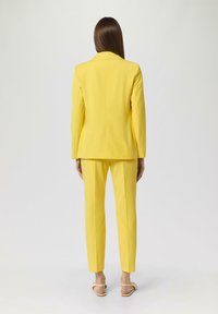 Completo giallo su misura con giacca strutturata e pantaloni slim-fit. Tessuto liscio, dettagli minimi e sandali con tacco blocco in un colore neutro.