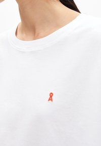 Biała bawełniana koszulka z okrągłym dekoltem i małym czerwonym haftowanym logo po lewej stronie klatki piersiowej. Miękka tekstura, casualowy design.