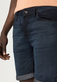 Dark denim shorts featuring a button closure, rolled cuffs in contrasting gray, and a slim fit design. Material has slight texture.