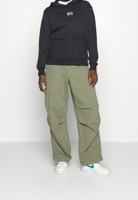 Svart hoodie med framfickor, kombinerad med lösa olivgrön byxor och vita sneakers med en grön swoosh. Avslappnad streetwear-stil.