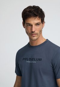 Grå bomullströja med rund halsringning, med en framtill broderad logotyp "POLO CLUB" och "ETABLERAD 2012" i kontrasterande färg.