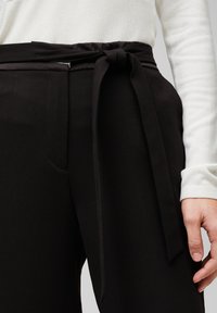 Svarta höga byxor tillverkade av ett slätt tyg, med en knuten slips vid midjan och sidofickor. Mjuk vit långärmad topp synlig.