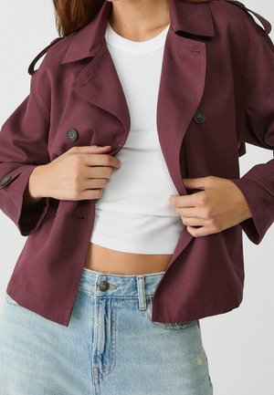 Personne ajustant une veste bordeaux boutonnée par-dessus un crop top blanc côtelé et un jean taille haute bleu clair avec un bouton visible.