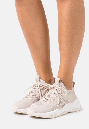 Béžové pletené tenisky se šněrováním a tlustými bílými podrážkami, nošené na bosých nohou na jednoduchém pozadí.