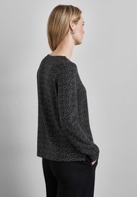 Schwarzes langärmeliges Oberteil mit weißem gepunktetem Muster, runder Ausschnitt, lockere Passform und seitlichem Taschen Detail, kombiniert mit schwarzen Hosen.