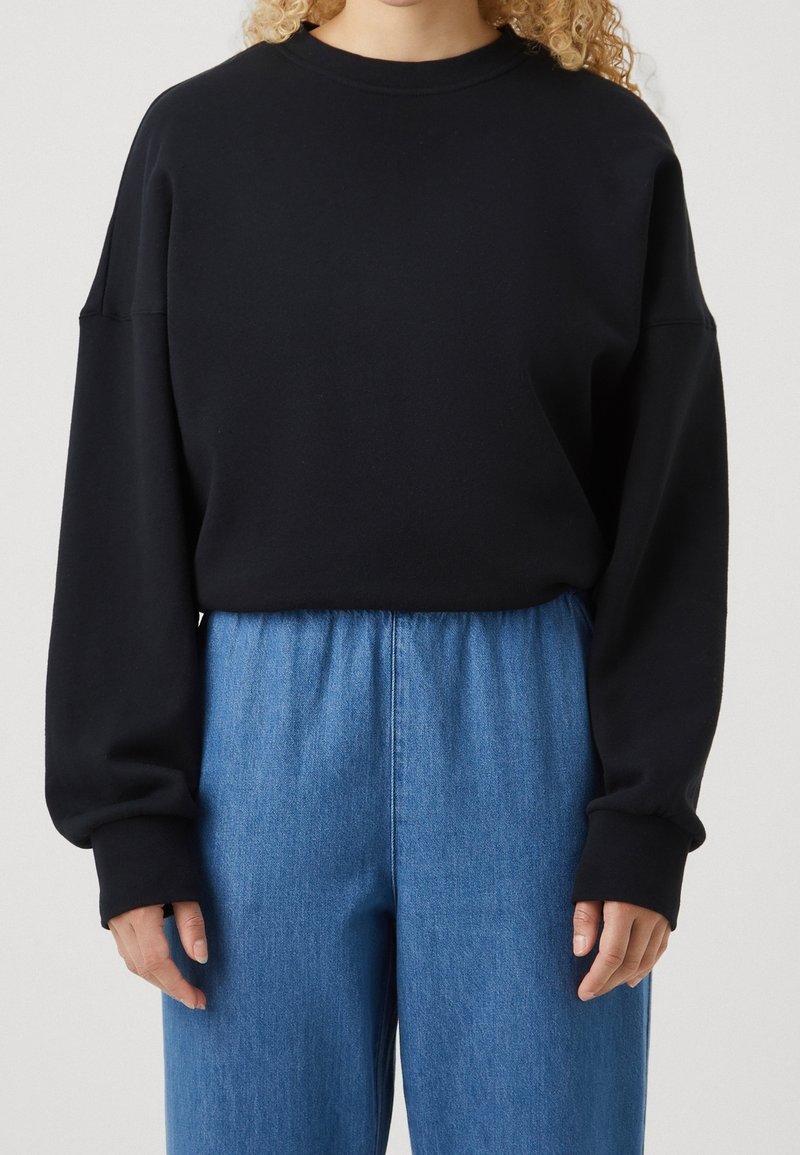 Personne aux cheveux bouclés blonds portant un haut noir ample à manches longues et un pantalon en denim bleu taille haute, sur un fond uni.