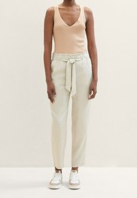 Ljusbeige ribbade tanktop, högmidjade ljusgröna byxor med knyt, och vita sneakers med beige detaljer. Neutral bakgrund.