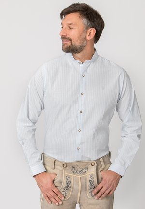 Mann in hellblauem Hemd mit Knöpfen und beigen, bestickten Lederhosen steht mit Händen in den Taschen und schaut seitlich.