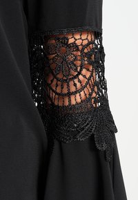 Svart klänningärm med intrikata svarta spetsdetaljer som visar blommönster med vågiga kanter och en slät tygtextur.