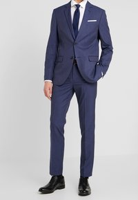 Námorně modrý oblek s texturovanou tkaninou, úzký střih, jednořadový kabát a klopové límce. Kombinovaný s bílou košilí a tmavými botami.